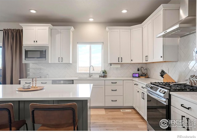 kitchen with stainless steel appliances, a sink, white cabinetry, light countertops, and wall chimney exhaust hood