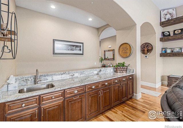 kitchen featuring baseboards, arched walkways, light wood-style flooring, light stone countertops, and a sink