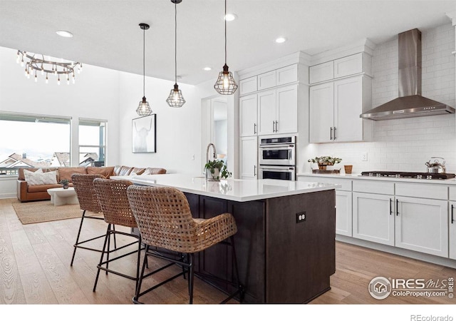 kitchen with gas stovetop, tasteful backsplash, double oven, light wood-style floors, and wall chimney exhaust hood