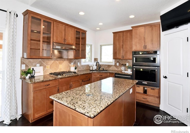 kitchen featuring glass insert cabinets, a center island, light stone countertops, under cabinet range hood, and stainless steel gas stovetop