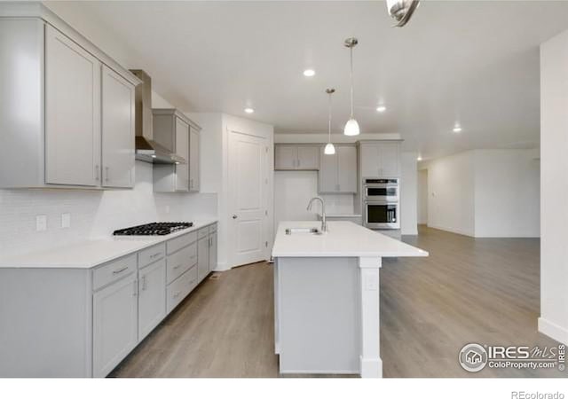 kitchen with gas stovetop, light countertops, a kitchen island with sink, a sink, and wall chimney exhaust hood