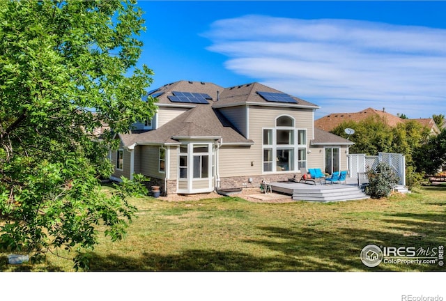 back of house with a shingled roof, a lawn, roof mounted solar panels, a deck, and stone siding