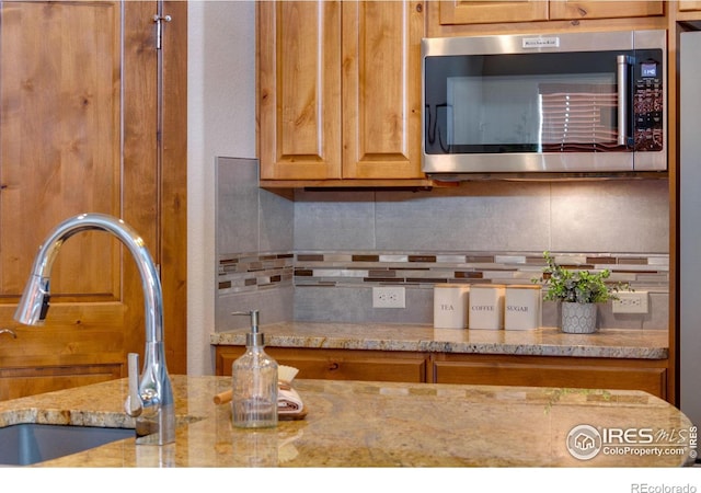 kitchen with stainless steel microwave, backsplash, brown cabinetry, a sink, and light stone countertops