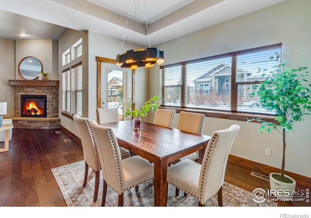 dining space featuring a stone fireplace, dark wood-type flooring, visible vents, baseboards, and a tray ceiling