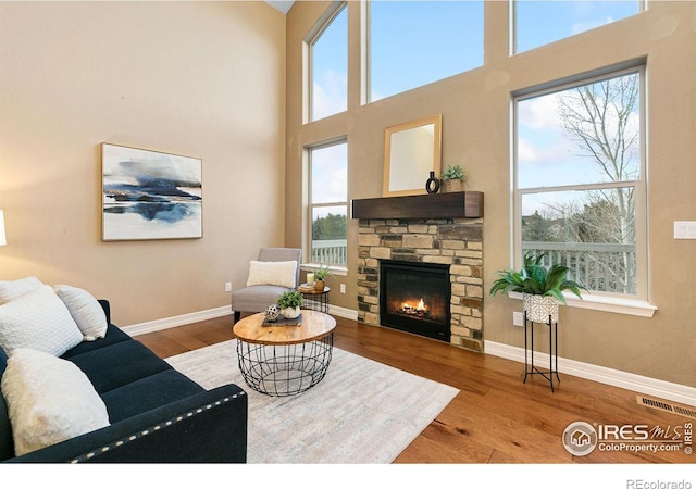 living room featuring baseboards, visible vents, a towering ceiling, wood finished floors, and a stone fireplace