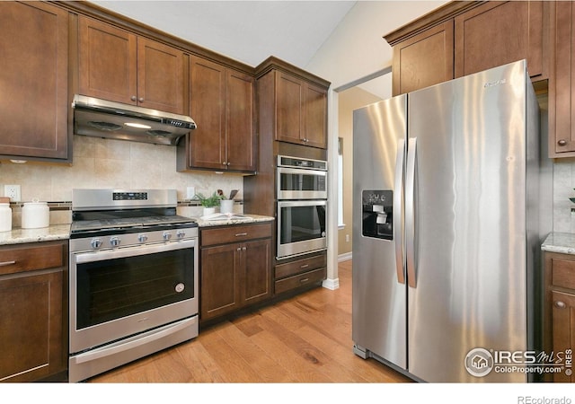 kitchen with under cabinet range hood, light wood-style floors, appliances with stainless steel finishes, backsplash, and light stone countertops