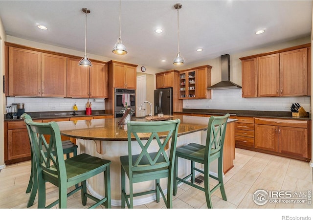 kitchen featuring a center island with sink, hanging light fixtures, appliances with stainless steel finishes, glass insert cabinets, and wall chimney exhaust hood