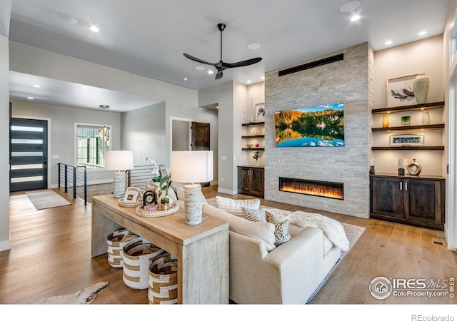 living area with light wood-style flooring, ceiling fan, a stone fireplace, built in shelves, and recessed lighting