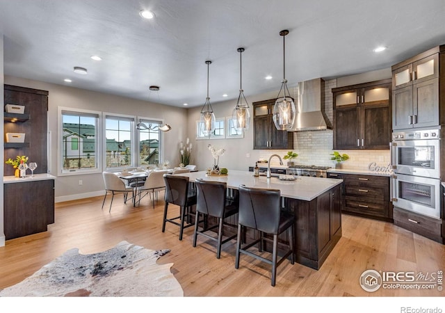 kitchen featuring decorative light fixtures, a center island with sink, glass insert cabinets, dark brown cabinets, and wall chimney exhaust hood