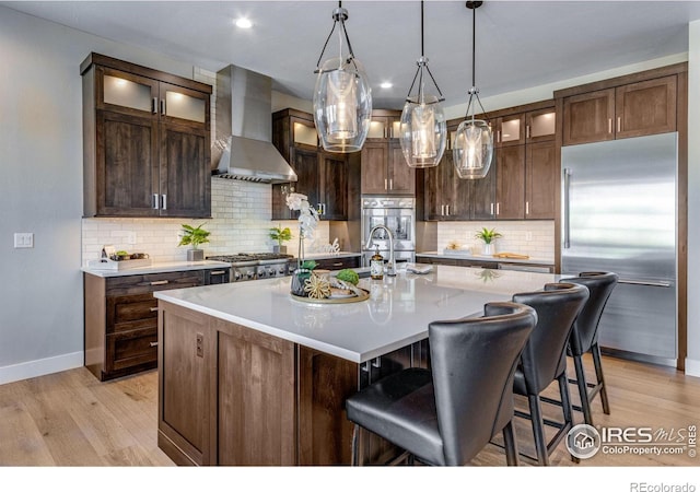 kitchen with stainless steel appliances, glass insert cabinets, an island with sink, and wall chimney exhaust hood