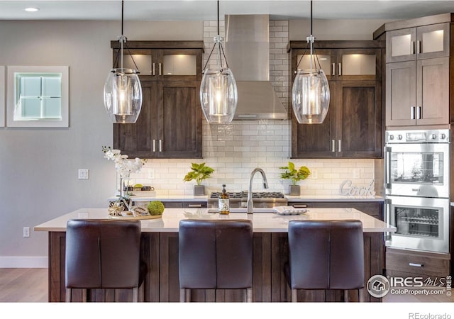 kitchen featuring dark brown cabinetry, light countertops, appliances with stainless steel finishes, wall chimney range hood, and glass insert cabinets