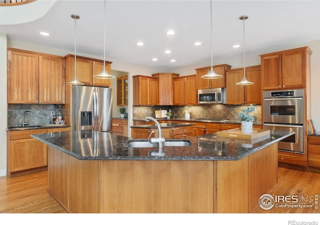 kitchen featuring a center island with sink, brown cabinetry, decorative light fixtures, stainless steel appliances, and a sink