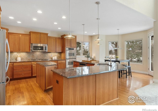 kitchen with a center island with sink, dark stone counters, appliances with stainless steel finishes, decorative light fixtures, and a sink