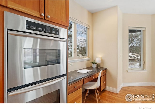 kitchen featuring double oven, brown cabinetry, dark stone counters, built in study area, and light wood-type flooring