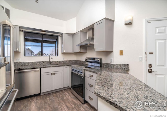 kitchen with wall chimney range hood, appliances with stainless steel finishes, gray cabinetry, a sink, and dark stone countertops