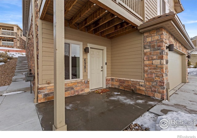 snow covered property entrance with a garage, stone siding, and a balcony