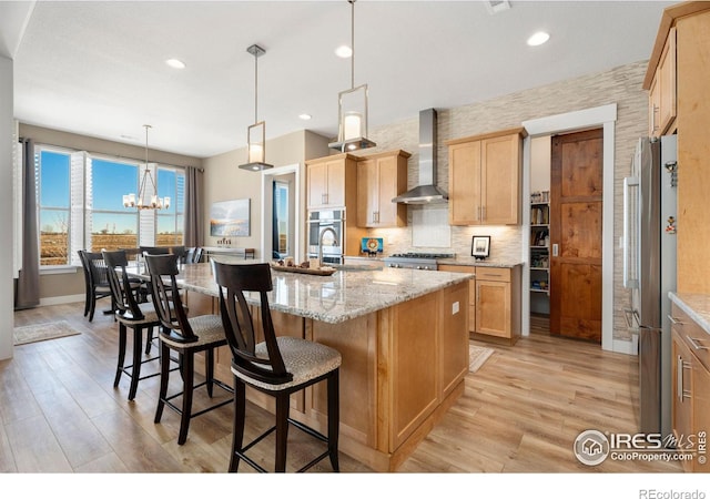 kitchen with wall chimney range hood, appliances with stainless steel finishes, light stone countertops, an island with sink, and pendant lighting