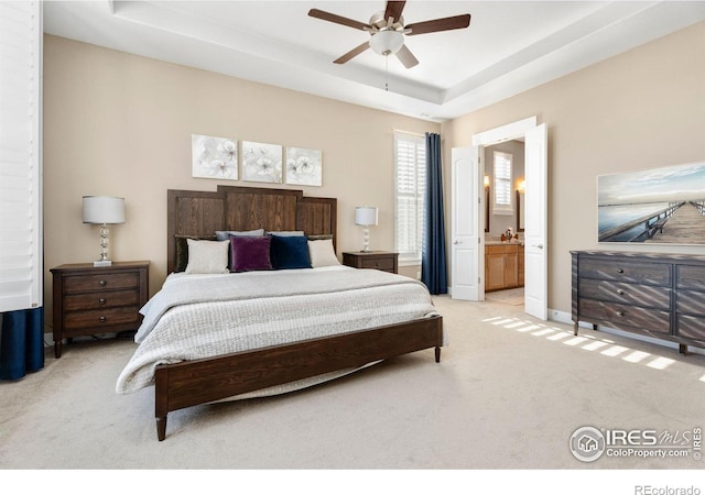 bedroom with ensuite bathroom, light colored carpet, a ceiling fan, baseboards, and a raised ceiling