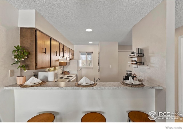 kitchen with a breakfast bar, light countertops, brown cabinetry, a peninsula, and under cabinet range hood