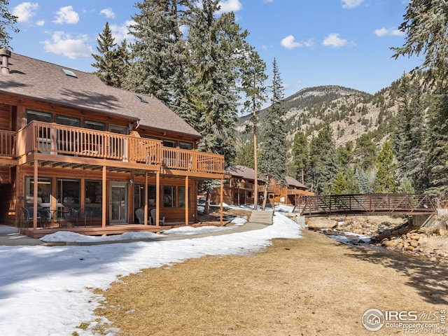 snow covered property with a shingled roof, a lawn, and a deck with mountain view