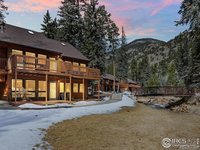 snow covered rear of property featuring a deck with mountain view, a yard, and a shingled roof