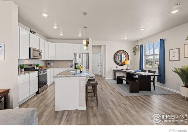 kitchen with light stone countertops, a kitchen island with sink, stainless steel appliances, white cabinetry, and pendant lighting