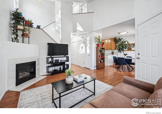 living room featuring a tiled fireplace, light wood-type flooring, a towering ceiling, and baseboards