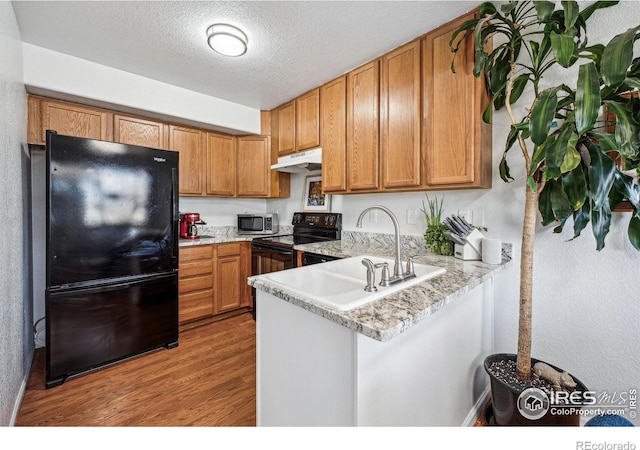 kitchen featuring brown cabinetry, a peninsula, under cabinet range hood, light wood-type flooring, and black appliances