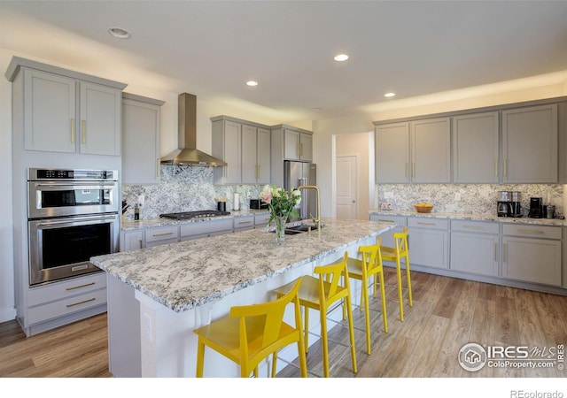 kitchen featuring wall chimney exhaust hood, light stone counters, stainless steel appliances, gray cabinetry, and a sink