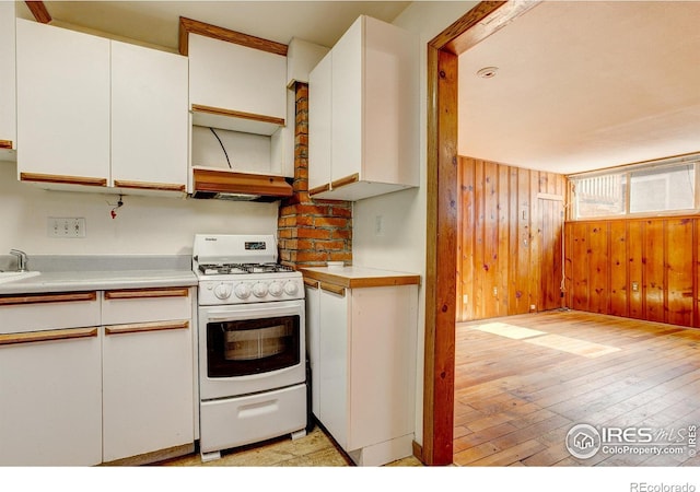 kitchen featuring light wood finished floors, white gas range, light countertops, white cabinets, and wood walls