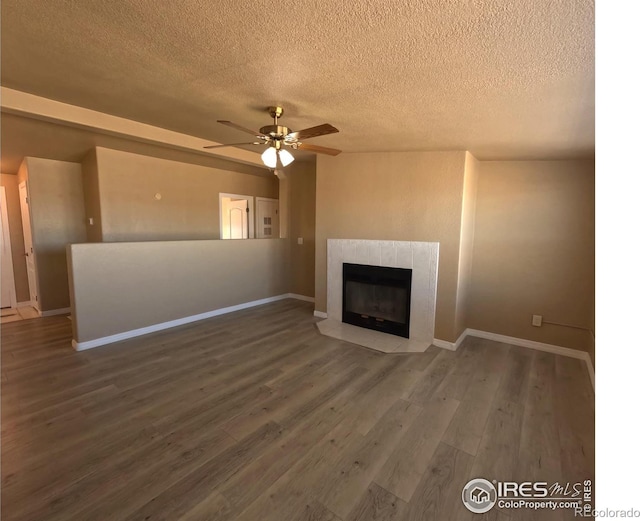 unfurnished living room with a tiled fireplace, ceiling fan, a textured ceiling, wood finished floors, and baseboards