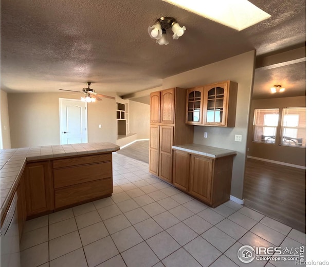 kitchen featuring tile countertops, light tile patterned floors, a ceiling fan, dishwasher, and glass insert cabinets