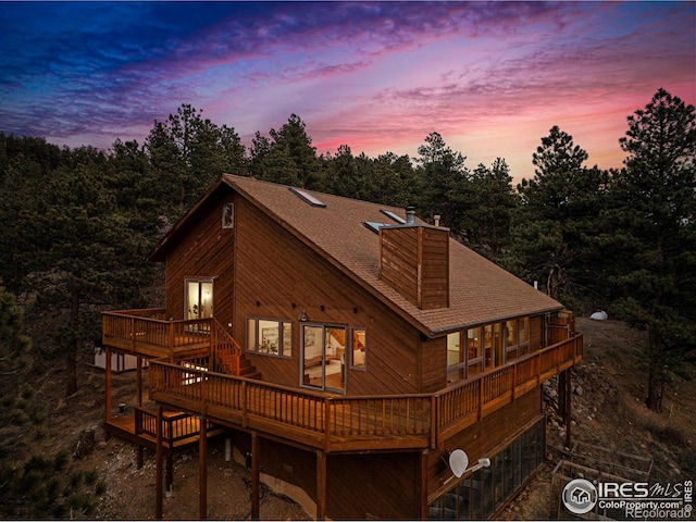 back of house at dusk featuring a shingled roof, a chimney, and a wooden deck