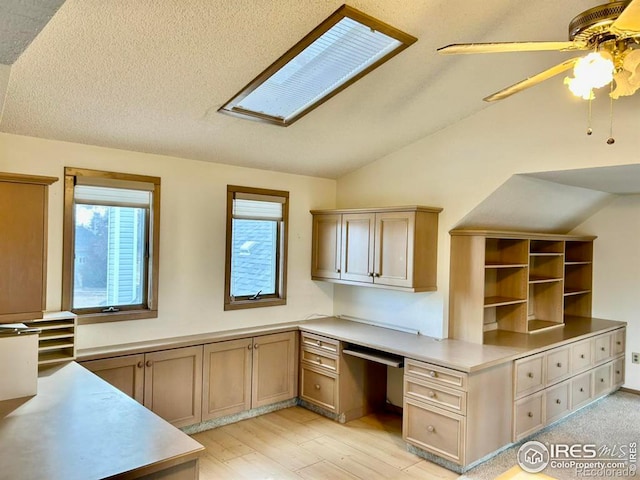 unfurnished office with light wood finished floors, vaulted ceiling with skylight, ceiling fan, a textured ceiling, and built in desk