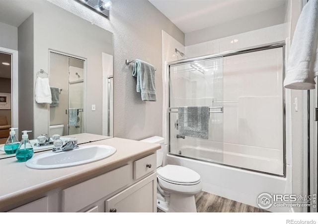 bathroom with a textured wall, toilet, shower / bath combination with glass door, wood finished floors, and vanity