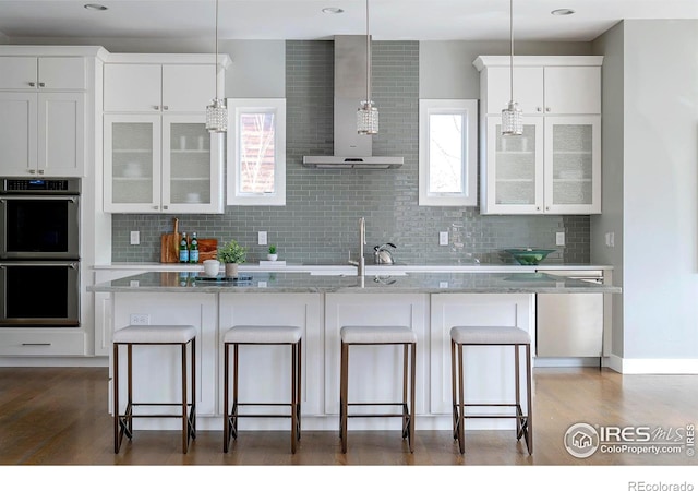 kitchen with double oven, wall chimney range hood, glass insert cabinets, and a breakfast bar