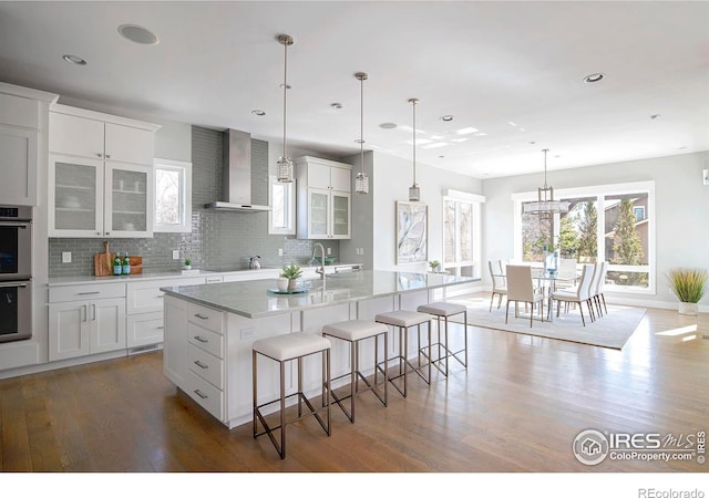 kitchen with glass insert cabinets, wall chimney range hood, a kitchen breakfast bar, and wood finished floors