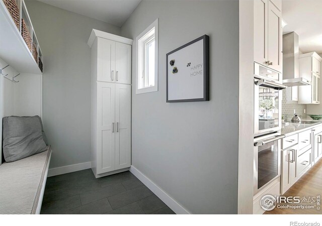 interior space with tasteful backsplash, baseboards, light countertops, wall chimney range hood, and white cabinetry