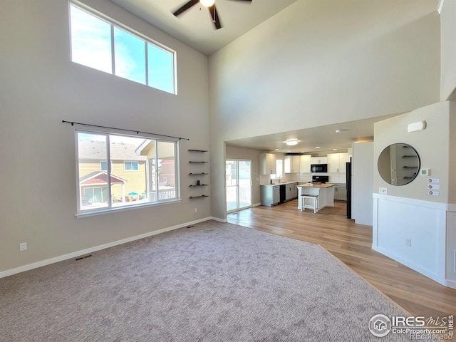 unfurnished living room with visible vents, ceiling fan, baseboards, light wood-style floors, and a towering ceiling