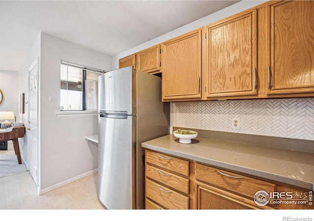 kitchen with light floors, backsplash, brown cabinetry, freestanding refrigerator, and baseboards