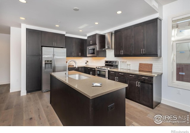 kitchen featuring a sink, appliances with stainless steel finishes, wall chimney range hood, decorative backsplash, and light wood finished floors