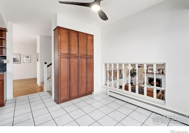 interior space featuring light tile patterned floors, a baseboard heating unit, a ceiling fan, a brick fireplace, and brown cabinets