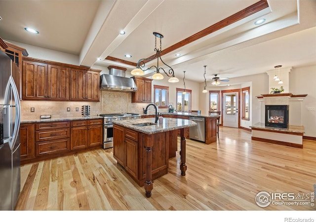 kitchen with a center island with sink, a glass covered fireplace, stainless steel appliances, wall chimney range hood, and a sink