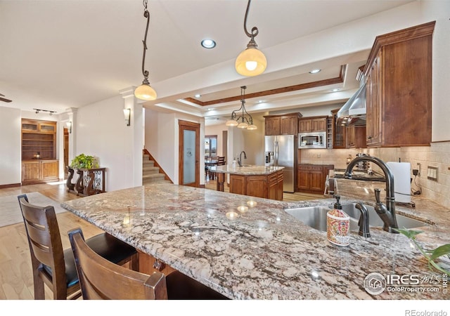 kitchen with a tray ceiling, brown cabinets, stainless steel appliances, light wood-style flooring, and light stone countertops