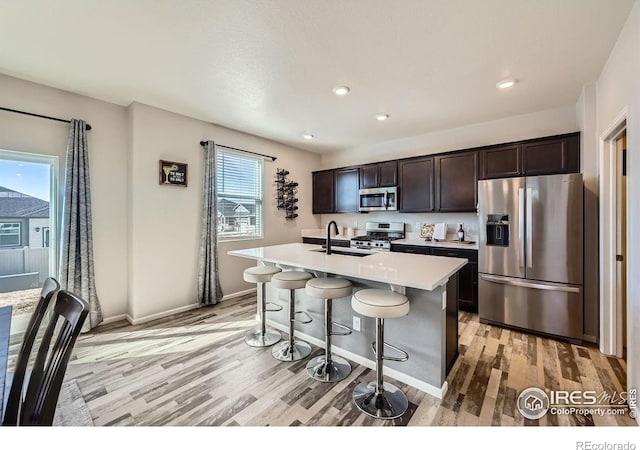 kitchen with stainless steel appliances, light wood-style flooring, a sink, an island with sink, and dark brown cabinets