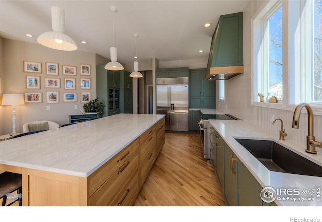 kitchen featuring decorative backsplash, high end appliances, green cabinetry, wall chimney range hood, and a sink