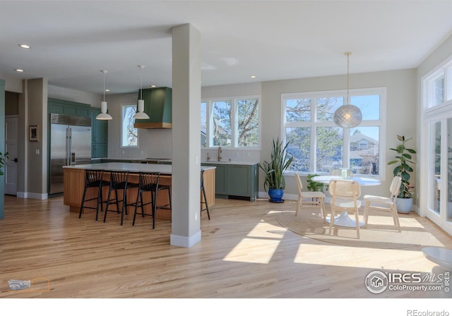 interior space with a sink, light wood-style floors, a kitchen breakfast bar, wall chimney range hood, and stainless steel built in refrigerator