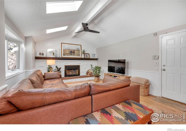 living room featuring a textured ceiling, vaulted ceiling with skylight, a wainscoted wall, light wood-style floors, and a glass covered fireplace