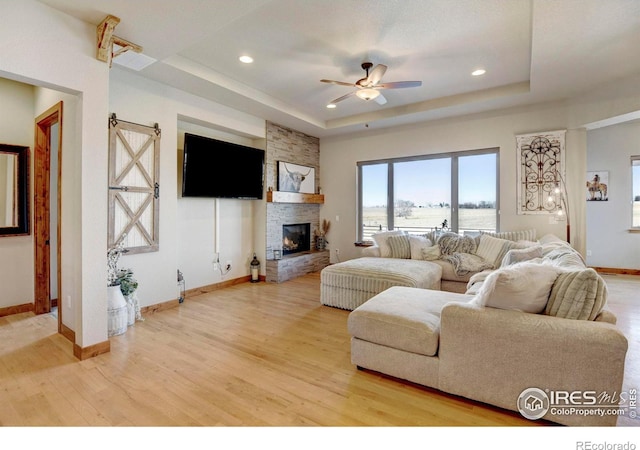 living area featuring baseboards, a stone fireplace, ceiling fan, a tray ceiling, and light wood-style flooring