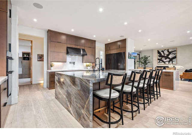 kitchen with decorative backsplash, light wood-style flooring, modern cabinets, wall chimney range hood, and black fridge